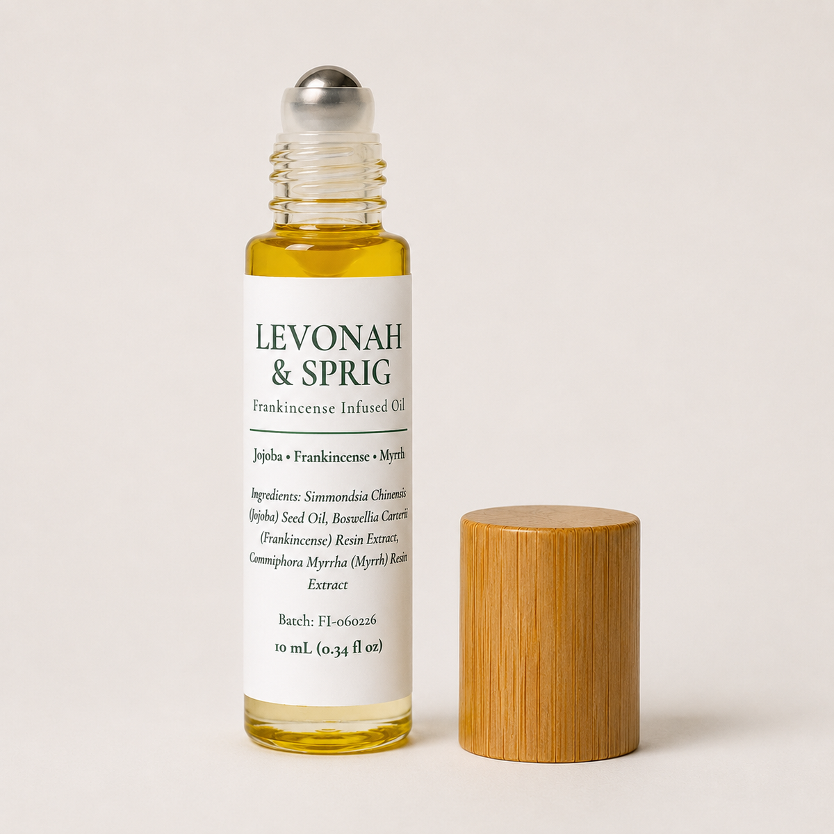 A clear glass bottle of Levonah & Sprig Frankincense Infused Oil with a white label and silver rollerball cap, shown next to a light wood cap on a warm off-white background.